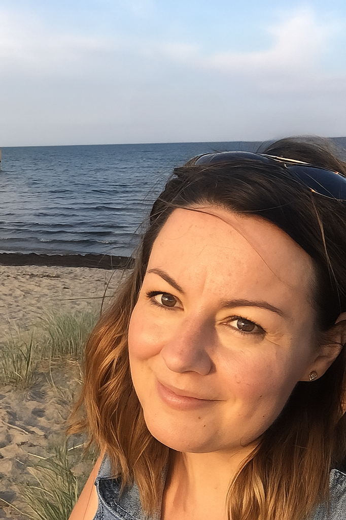 A woman with brown hair and sunglasses resting on her head, smiling at the camera, on a beach with sand, green grass, and ocean in the background under a partly cloudy sky.