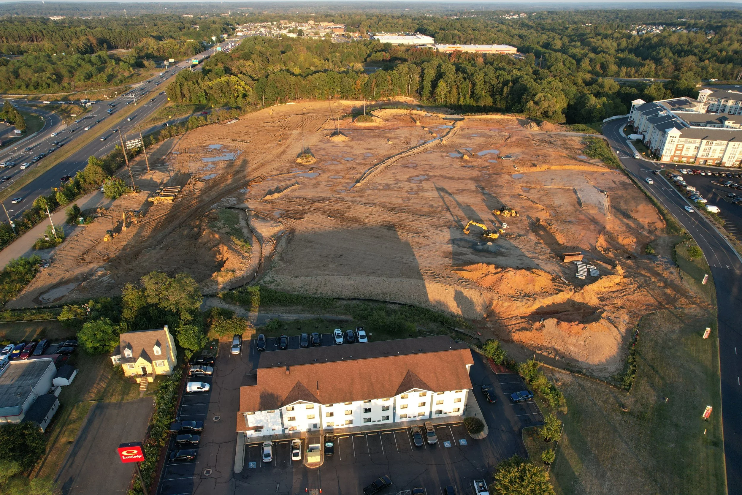 Aerial view of a construction site in an urban area with dirt and excavation equipment, surrounded by buildings, parking lots, and a highway.