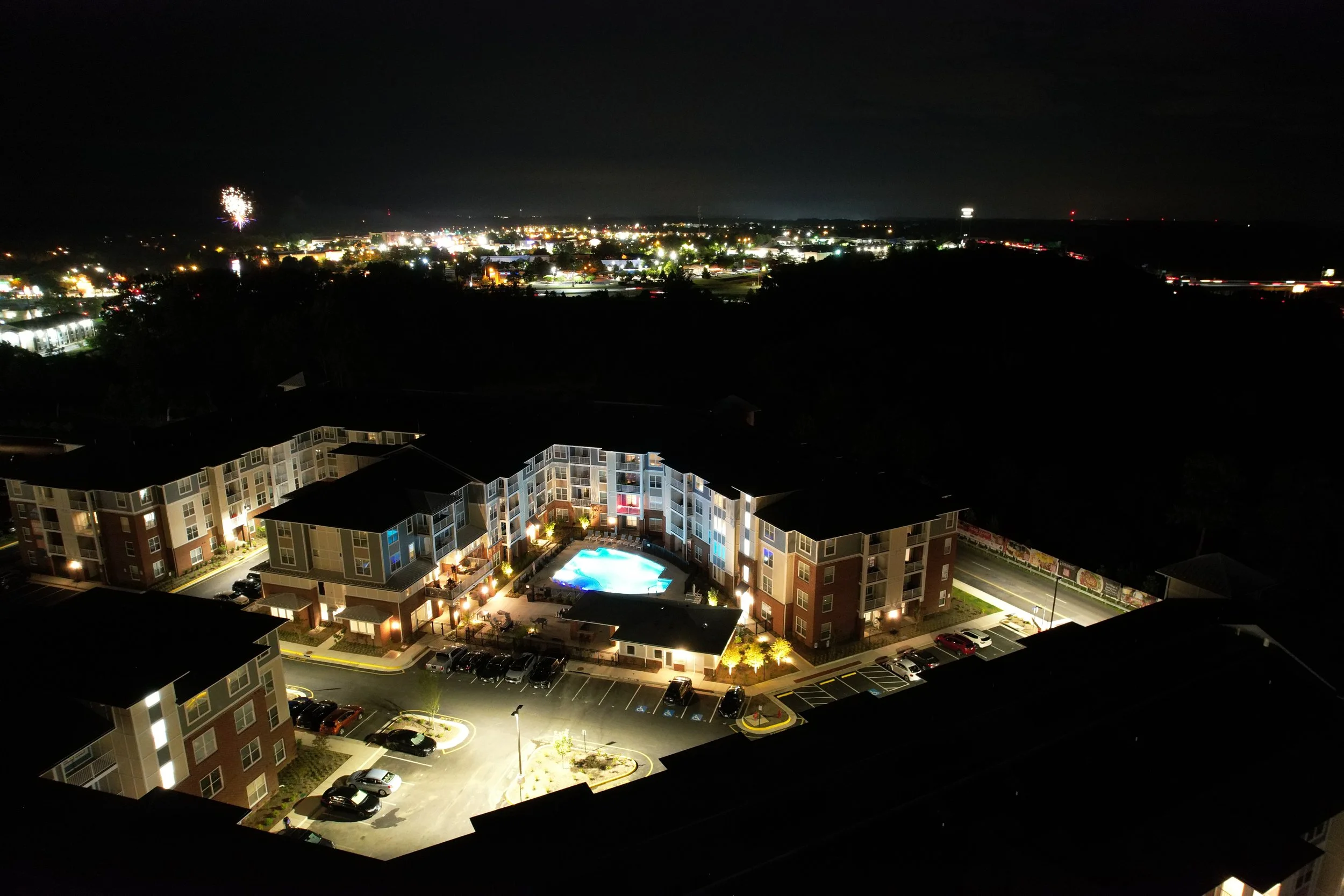 Night view of an apartment complex with a lit swimming pool, parking lot, and surrounding buildings under a dark sky with city lights and fireworks in the distance.