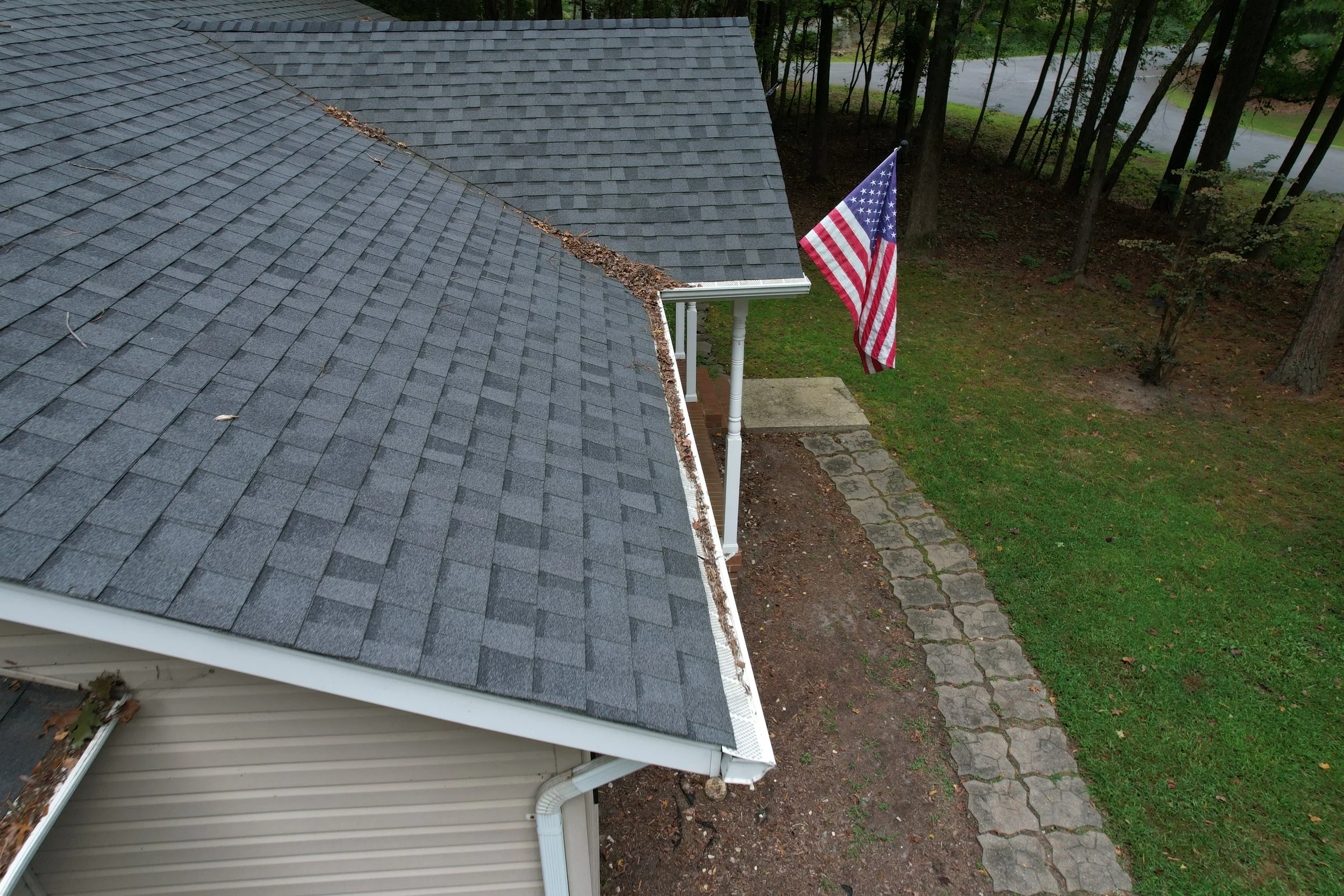 A house with a grey shingle roof, a white gutter, and a small white porch. An American flag is mounted on a pole near the porch, and there is a stone pathway leading away from the house. There are trees and grass in the background.