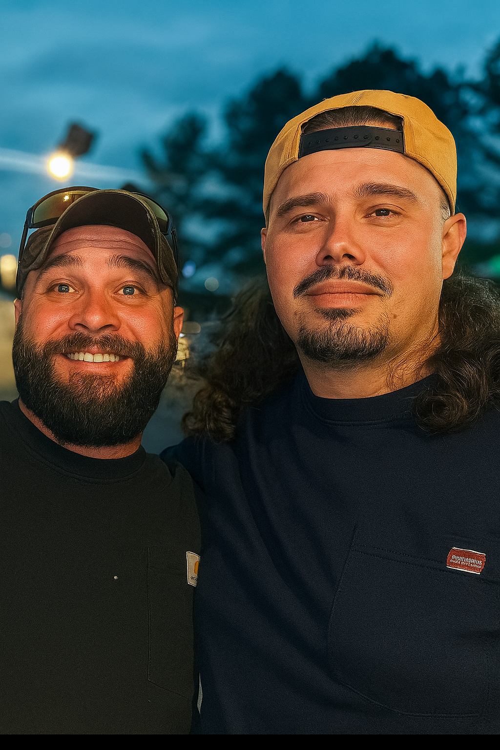 Two men smiling, standing close together outdoors during the evening, with a blurred background of trees and streetlights.