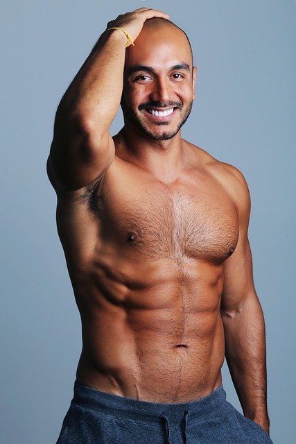 A shirtless man with a muscular build, short beard, and short hair smiling with one hand on his head against a plain light blue background.
