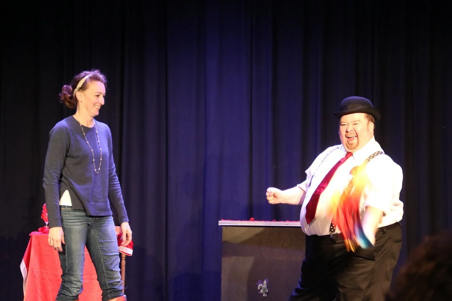 Two people on stage, a woman on the left smiling and a man on the right wearing a bowler hat, white shirt, red tie, and colorful suspenders, appearing to perform or act, with a black curtain backdrop.