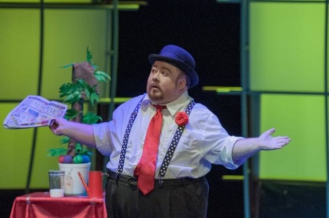 A performer on stage wearing a blue bowler hat, white shirt, red tie, suspenders, and a red flower pin, gesturing with arms outstretched in front of a bright yellow-green background.