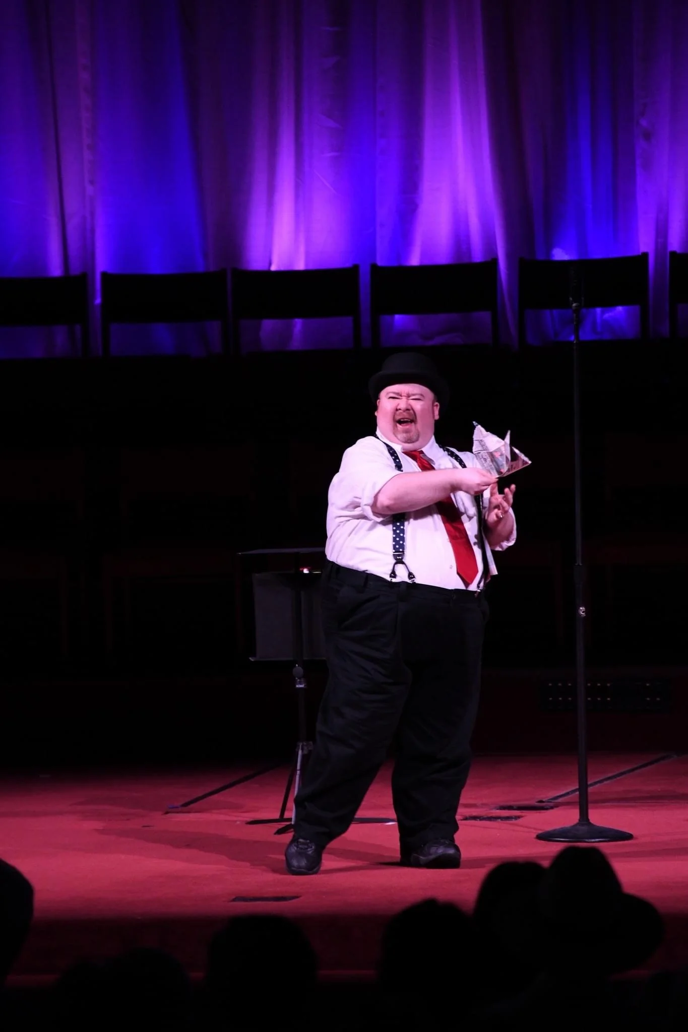 A male performer dressed as a clown or comedic character on stage, holding a paper origami, with a purple-lit curtain in the background and a microphone stand nearby.