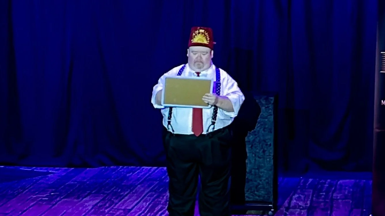 A man dressed in a white shirt, red tie, black pants, suspenders, and a red hat with a yellow decoration, standing on a stage with a dark curtain behind him, looking at a framed picture.