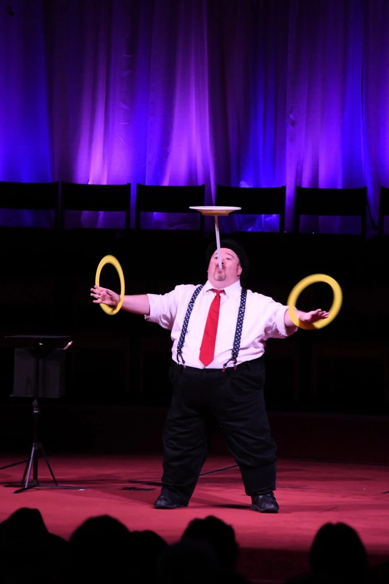 A circus performer balancing a platform on his nose while spinning yellow rings on his fingers on stage with purple curtains in the background.