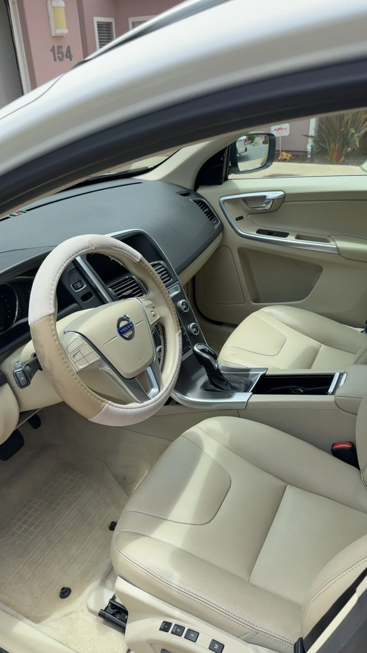Interior of a beige Volvo car with a leather steering wheel, beige seats, and center console, parked in front of a pink building with the number 154.