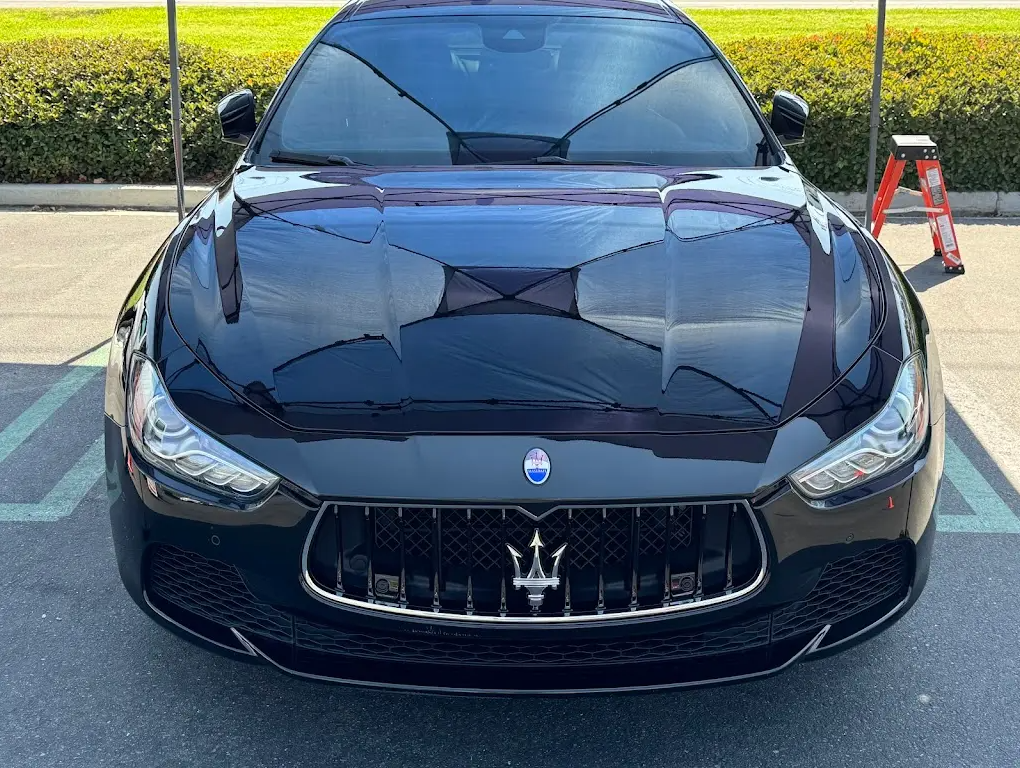Black Maserati sports car parked in a parking lot with hedge in the background.
