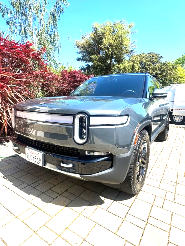 A gray electric SUV parked on a brick driveway, surrounded by trees and plants, with a clear blue sky in the background.