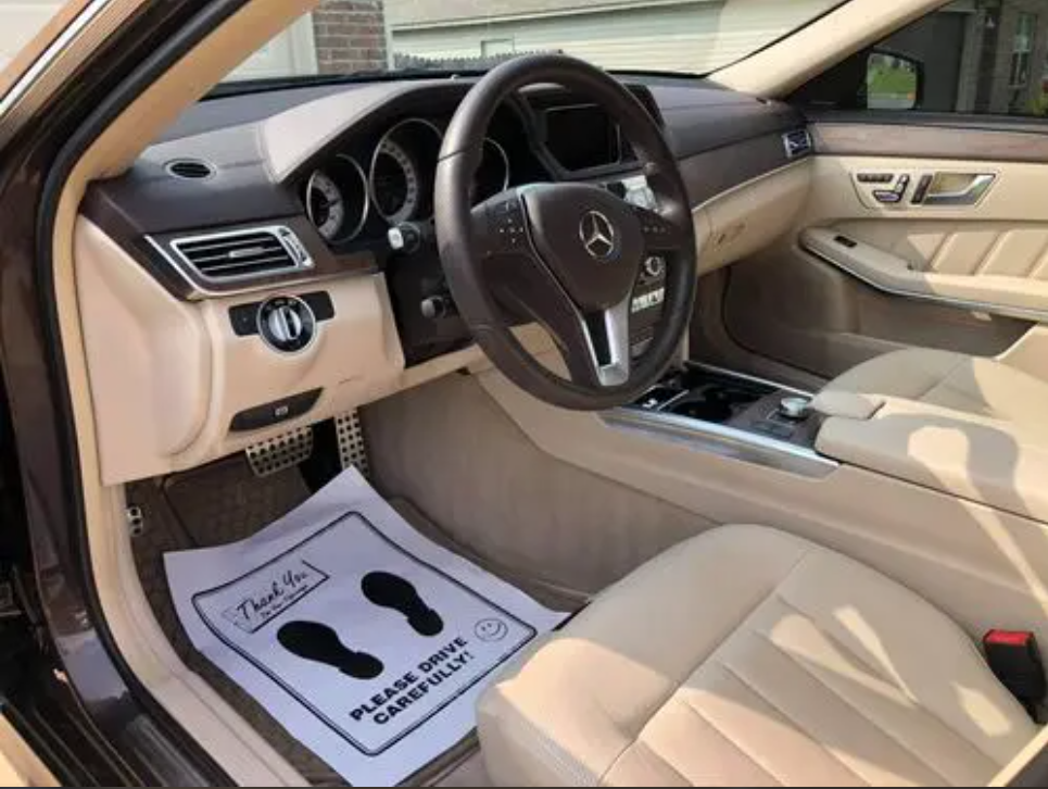 Interior of a luxury car with beige seats, steering wheel with Mercedes-Benz logo, center console, and floor mat that says 'Thank You! Please Drive Carefully'.
