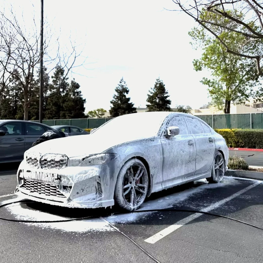 A white sedan covered in foam at a car wash, with soap and water on the surface, in a parking lot with trees and other cars in the background.