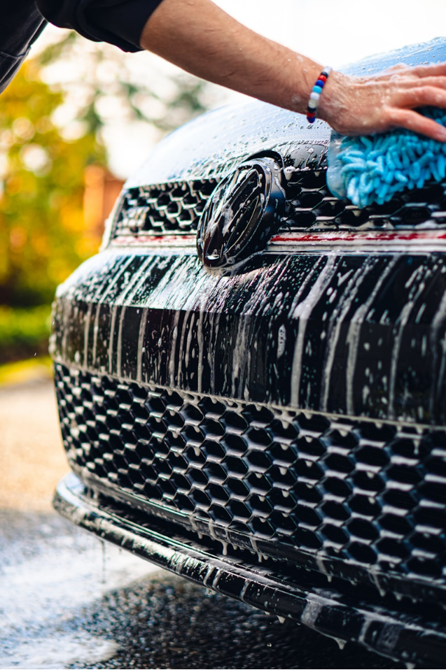 Person washing a black Volkswagen car with a blue sponge, with soap suds covering the front grille and hood.