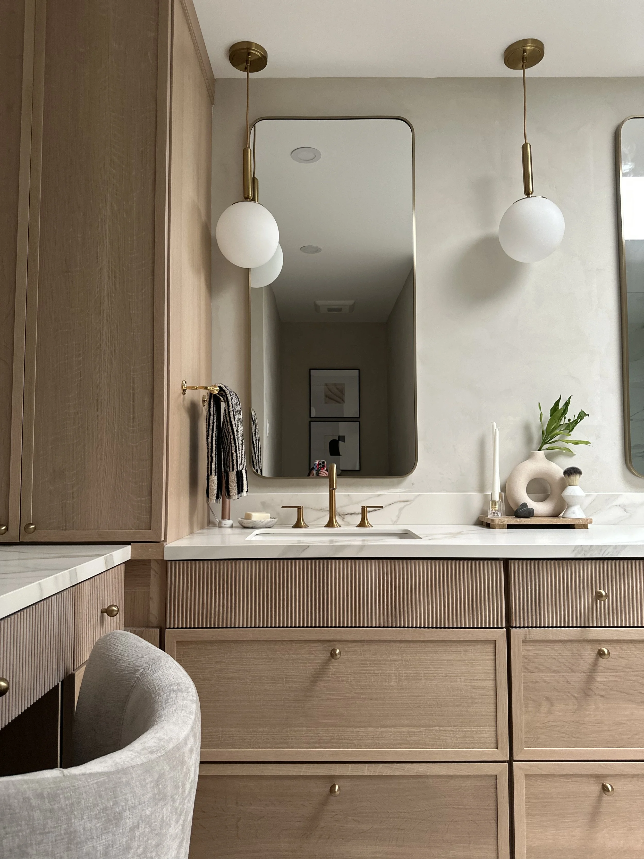A bathroom vanity with a wooden cabinet and a cream-colored marble countertop, a large rectangular mirror, two pendant lights with white globes, a gold faucet, and decorative items such as a vase with greenery and art prints on the wall.