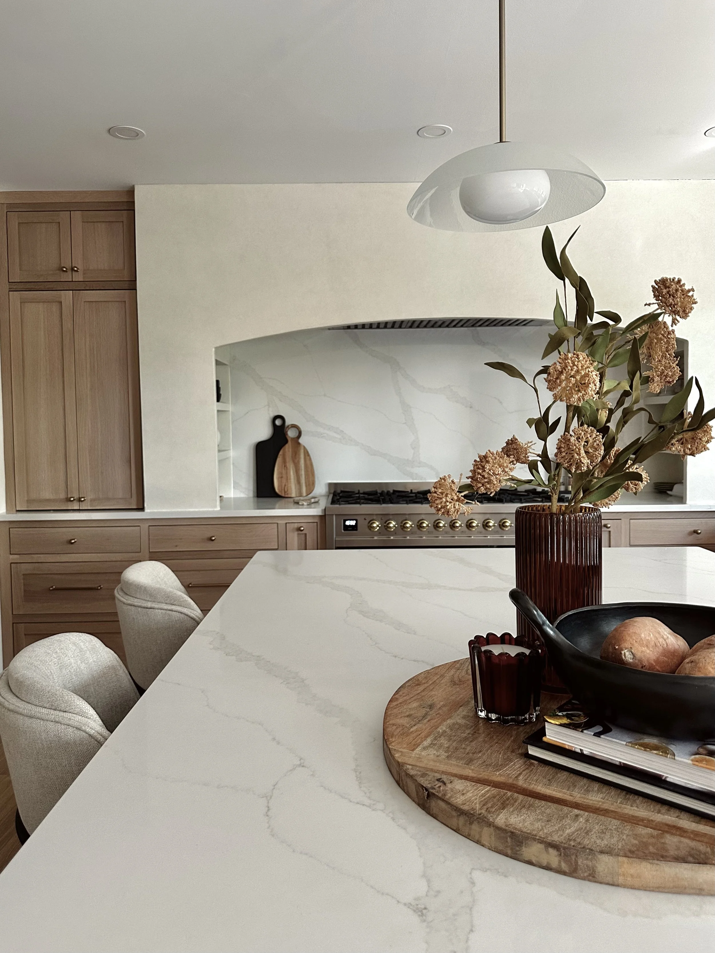 A modern kitchen with a white marble countertop, light wood cabinets, a decorative vase with dried flowers, and a black frying pan with a sweet potato on a wooden tray. There are white upholstered chairs around the table, and a contemporary white pendant light hanging from the ceiling.