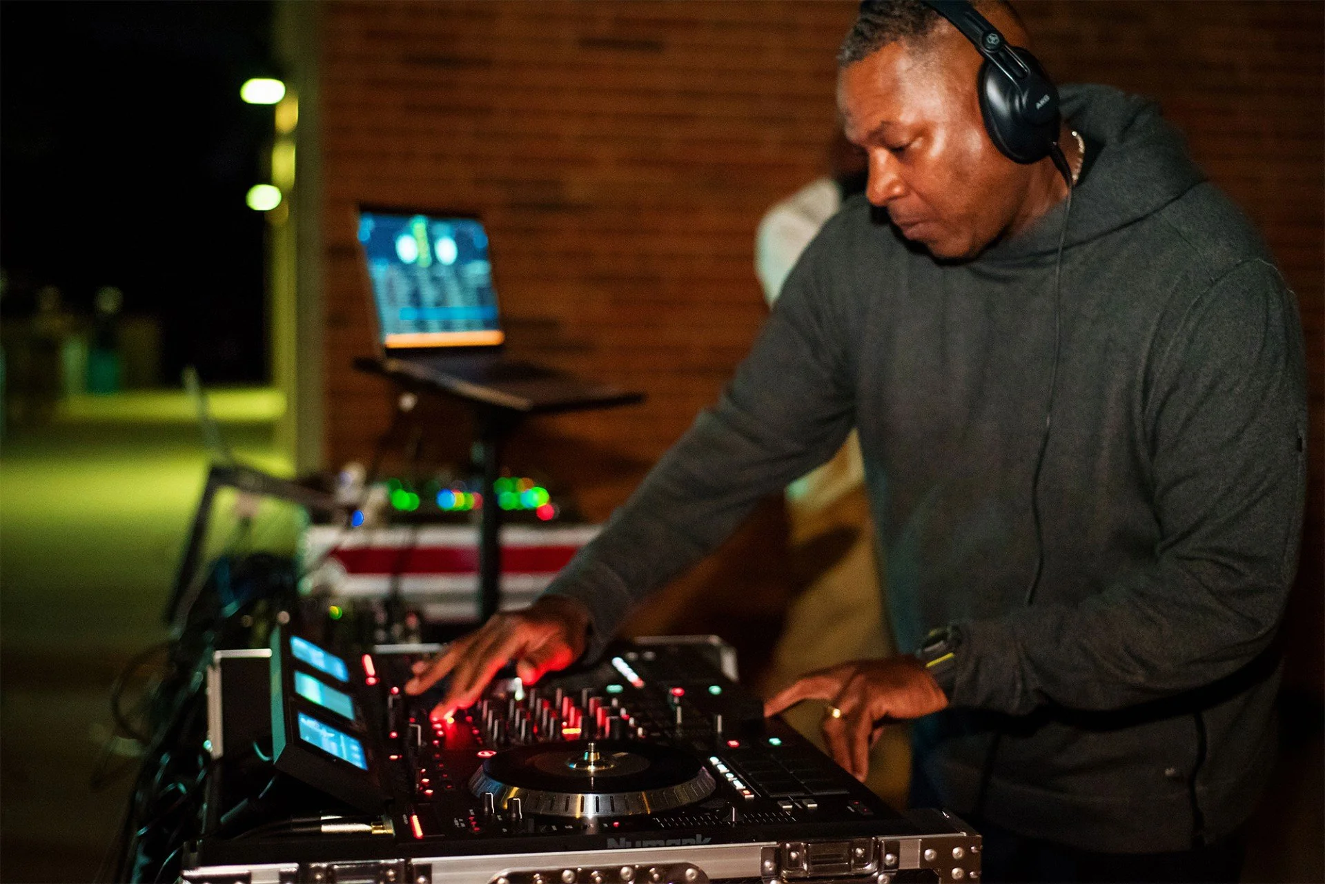 A man DJing at an indoor event, mixing music with professional DJ equipment, wearing headphones and a dark hoodie.