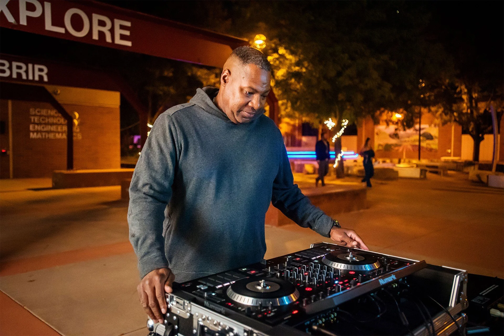 A man in a dark hoodie DJing outdoors at night, with trees and people in the background, illuminated by warm lighting.