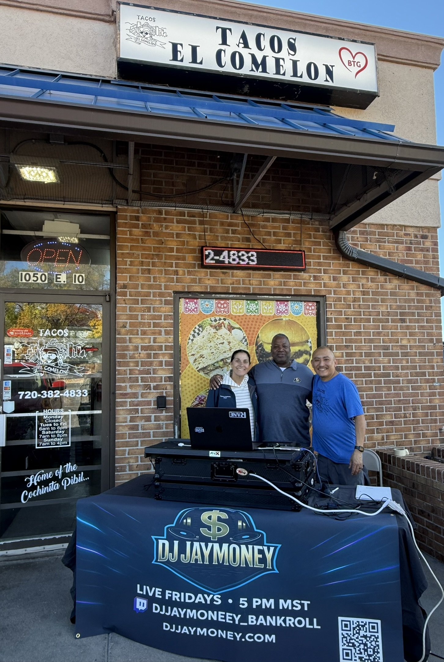 People standing behind a DJ booth outside a Mexican restaurant called Tacos El Comelon. The DJ banner reads DJ Jay Money, with streaming info and a QR code. The restaurant sign is above, and there is a digital signboard showing the time and temperatu