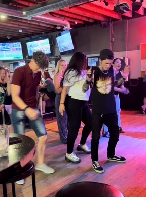 Group of young people dancing and having fun at a bar or nightclub with multiple TV screens in the background.