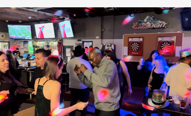 People dancing and socializing in a bar with multiple TV screens, dartboards, and colorful lighting.