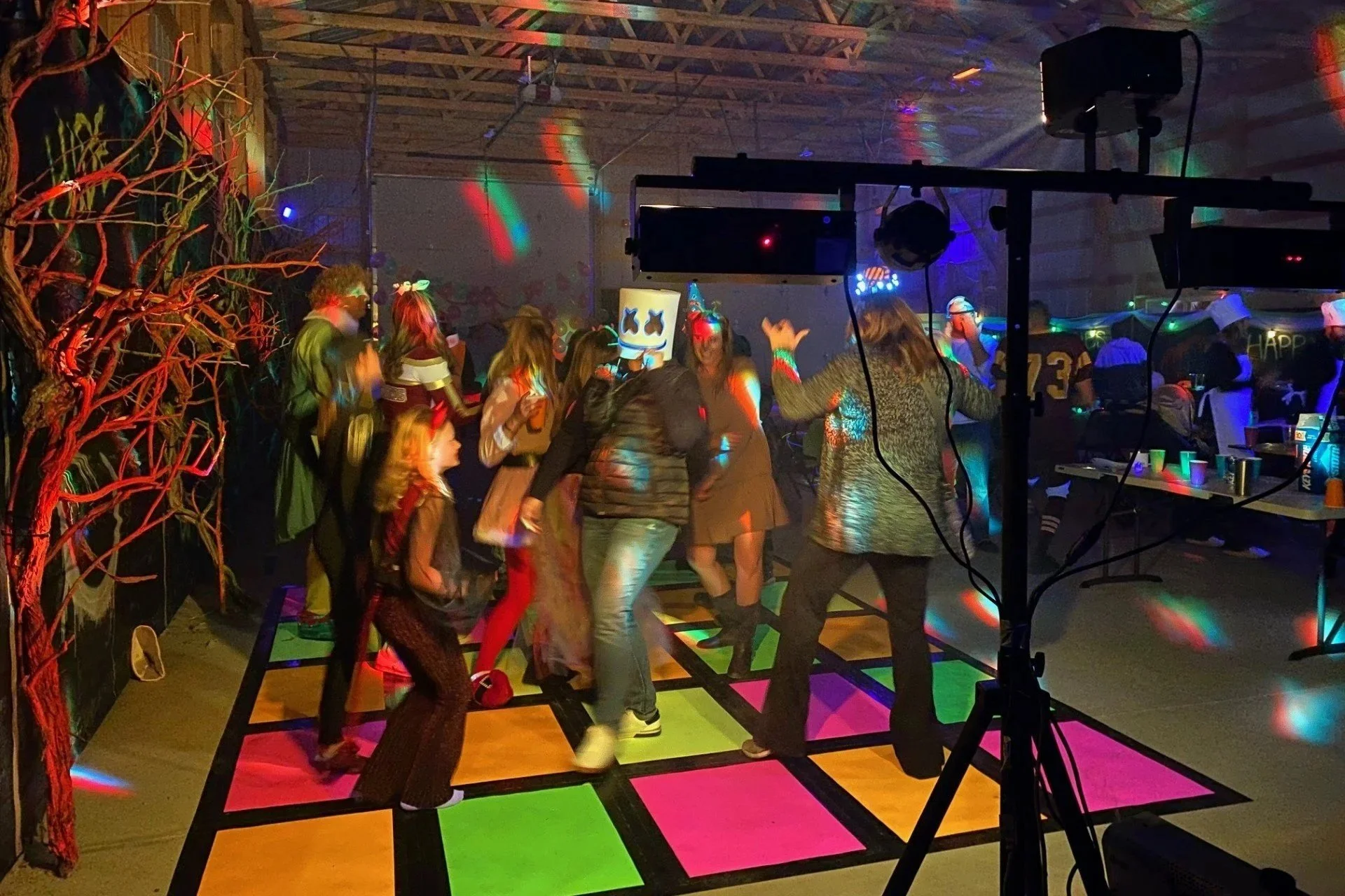 People dancing on a colorful illuminated dance floor at a party or celebration, with party lights and a DJ setup visible.