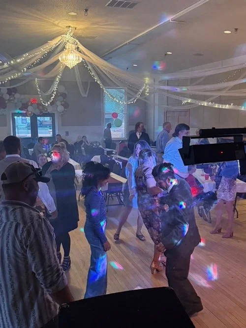 People dancing on a decorated ballroom floor with string lights and a chandelier, colorful disco lights, and windows with balloons outside.