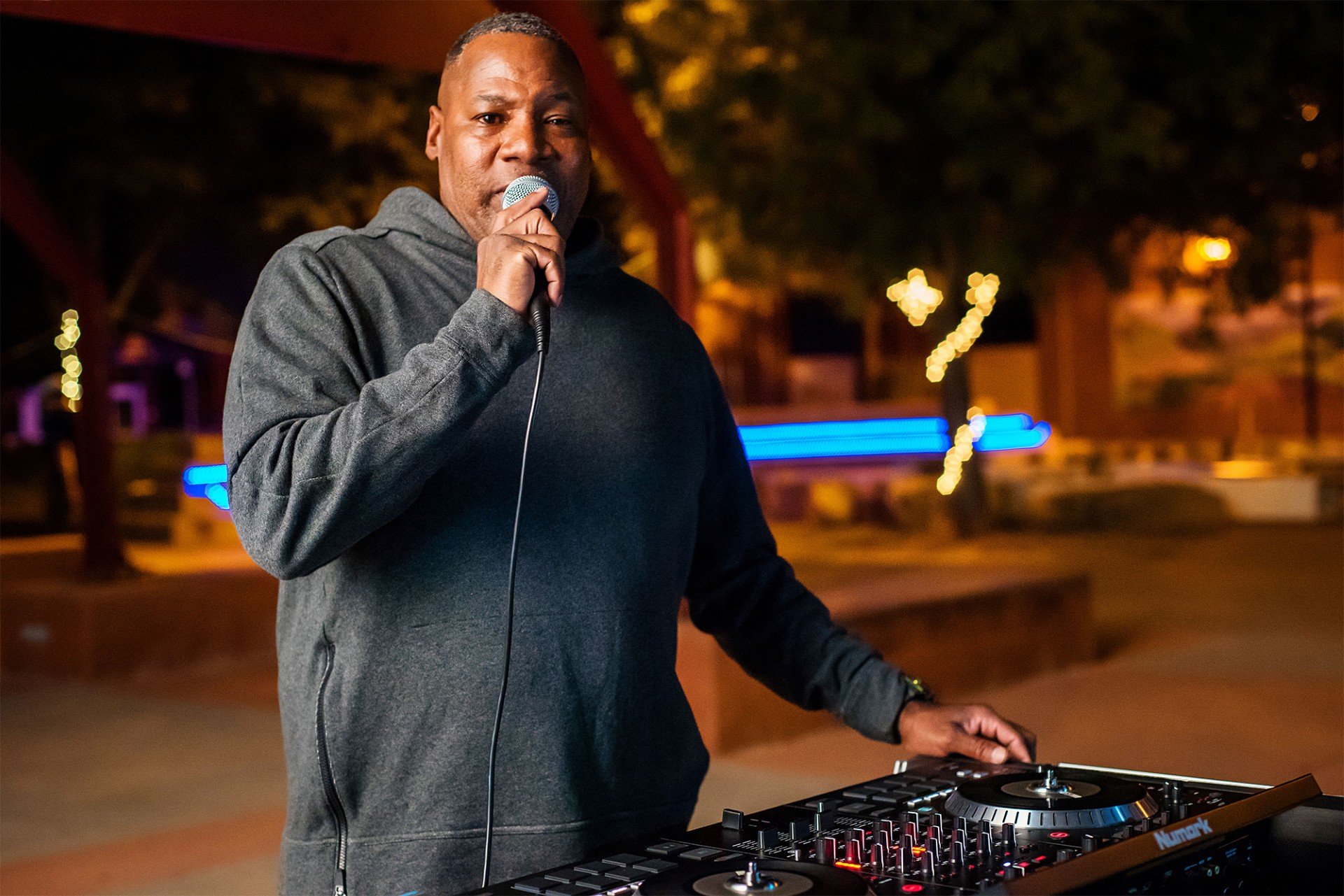 A man wearing a gray hoodie is holding a microphone and standing behind a DJ turntable, performing outdoors at night.