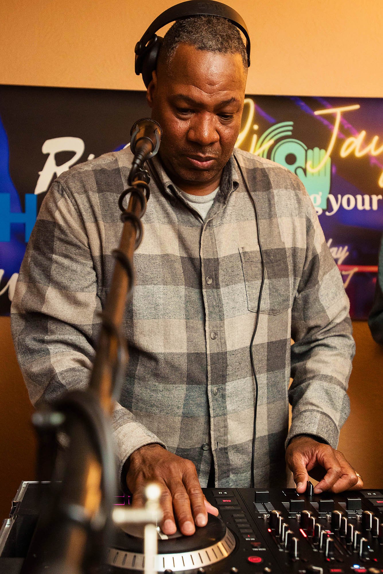 A man with short dark hair playing music on a DJ controller, wearing headphones, in a room with a colorful sign in the background.