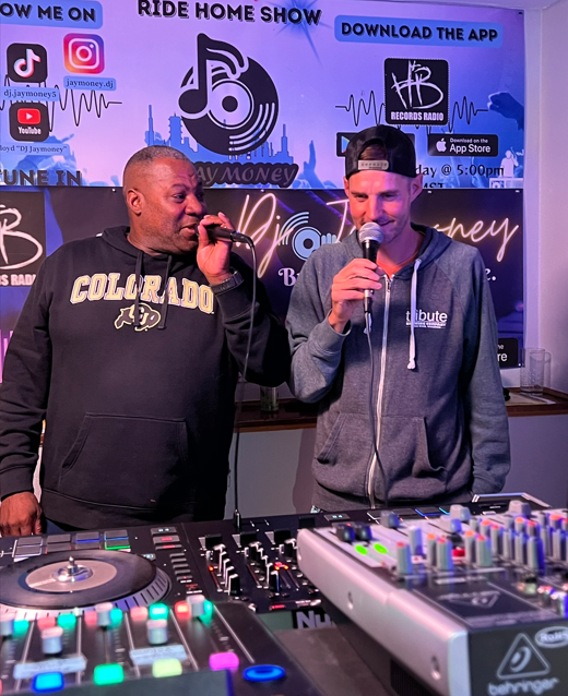 Two men are standing behind a DJ mixer with microphones, in a radio station studio. The background features blue and purple signs with radio station information, social media icons, and a city skyline graphic. One man wears a black hoodie with 'Color