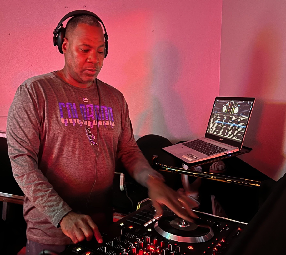 A DJ wearing a Colorado Rockies shirt and headphones is working with a DJ controller, with a laptop displaying music software nearby in a dimly lit room with pink lighting.