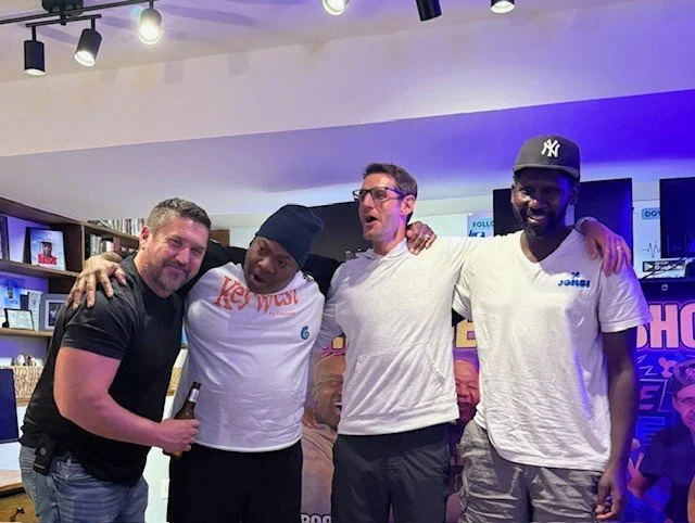 Four men standing together in a room, smiling, with a purple and blue background, and bookshelves with framed photos on the wall behind them.