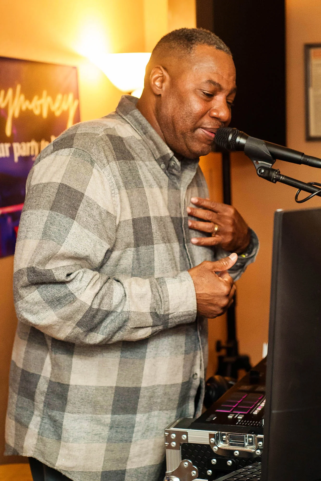 A man singing or speaking into a microphone while standing at a DJ setup, wearing a gray and white plaid shirt, in a warmly lit indoor setting.