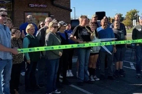 Group of people gathered outdoors for a ribbon-cutting ceremony, with a man holding large scissors, standing in front of a yellow ribbon.