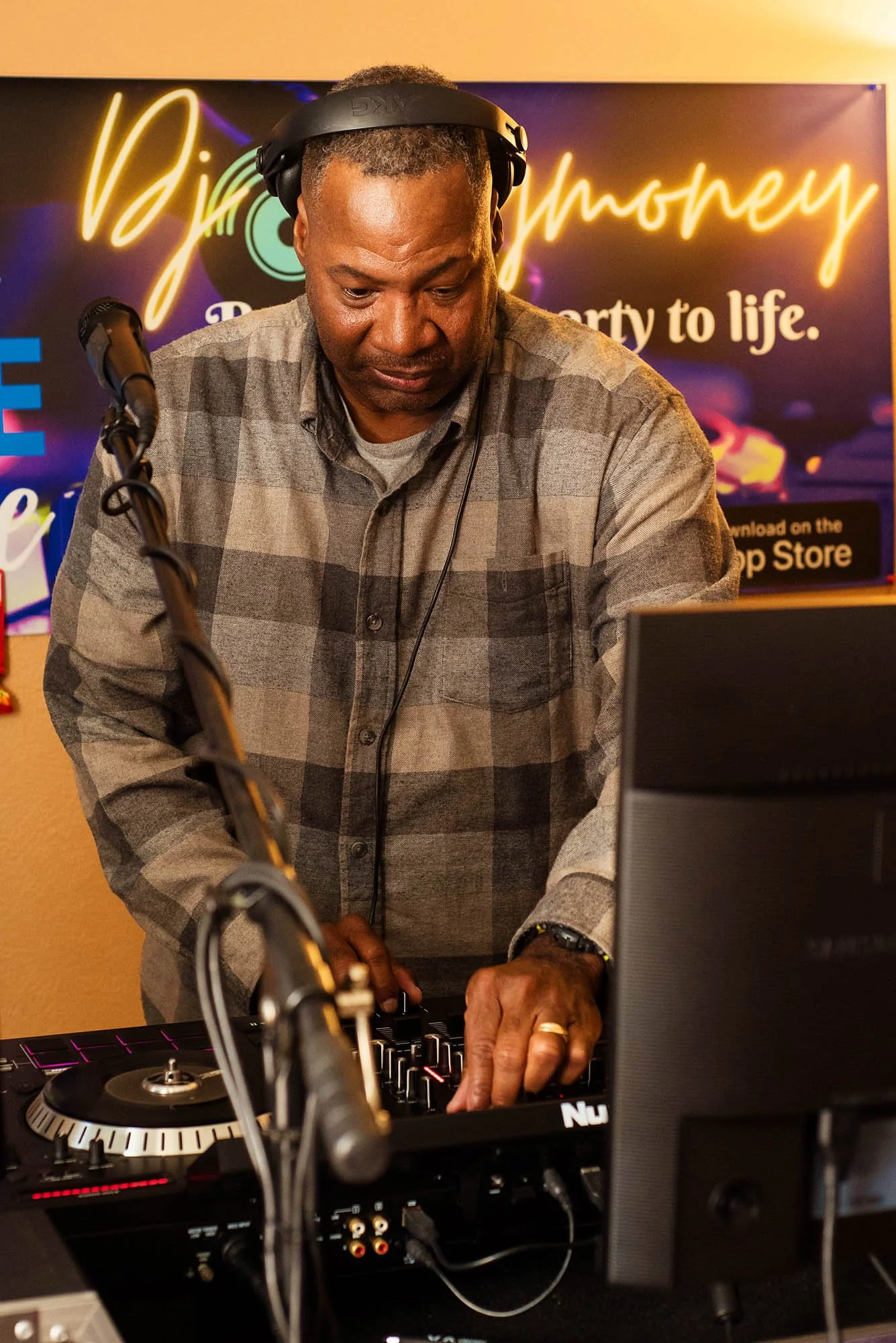 A man with gray hair and a beard wearing a gray plaid shirt and headphones DJing at a booth. He is adjusting controls on a DJ mixer in front of a colorful neon sign background.