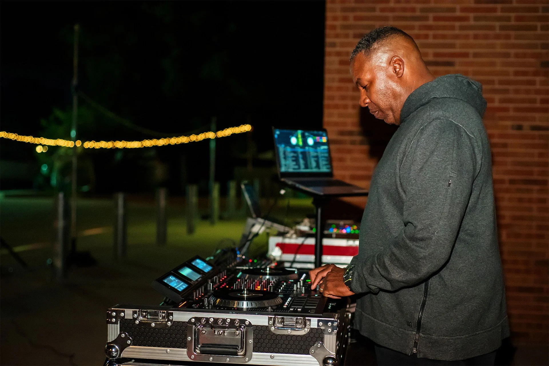 A DJ wearing a dark hoodie and black pants performing outdoors at night, operating a DJ mixer and laptop with colorful lights, with a brick wall behind him and string lights in the background.