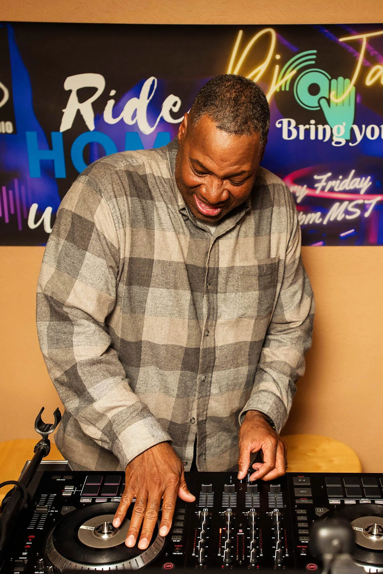 A man wearing a gray plaid shirt is DJing with a professional DJ controller, with neon signs and a promotional poster in the background.