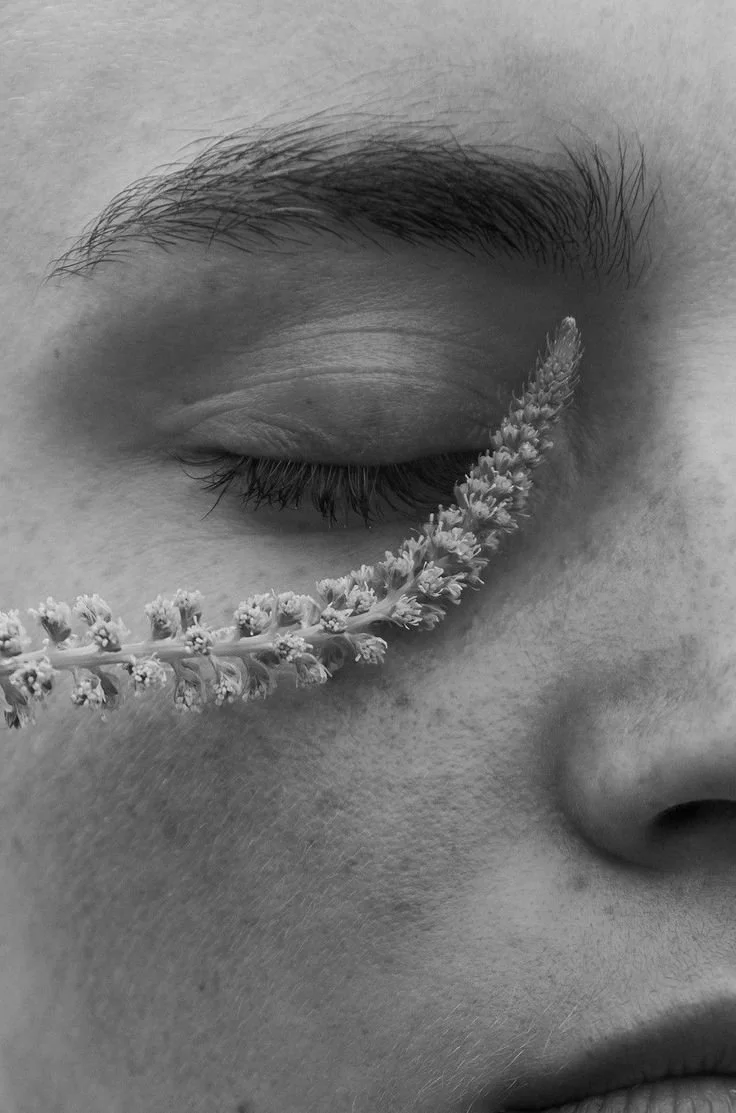 Close-up de uma pessoa com os olhos fechados, com uma flor de lavanda repousando na bochecha, em uma foto em preto e branco.