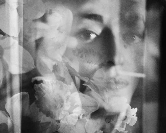 Retrato em preto e branco de uma mulher com flores e uma pessoa ao fundo