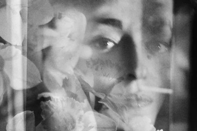 Retrato de uma pessoa com um sorriso suave, visto parcialmente através de uma superfície transparente com flores.