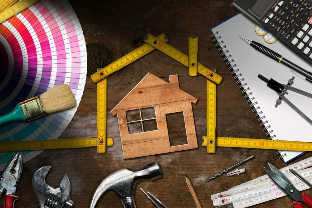 A wooden house model in the center surrounded by a tape measure, painting tools, and drafting tools on a wooden table.