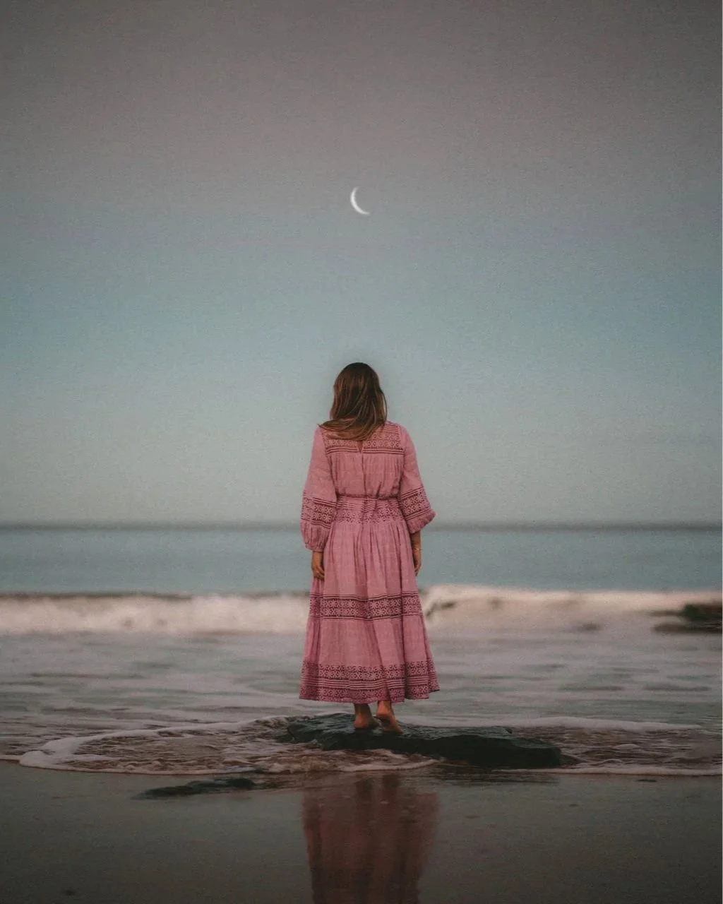 A woman in a pink boho dress standing on the shore, facing the ocean with a crescent moon visible in the serene sky.