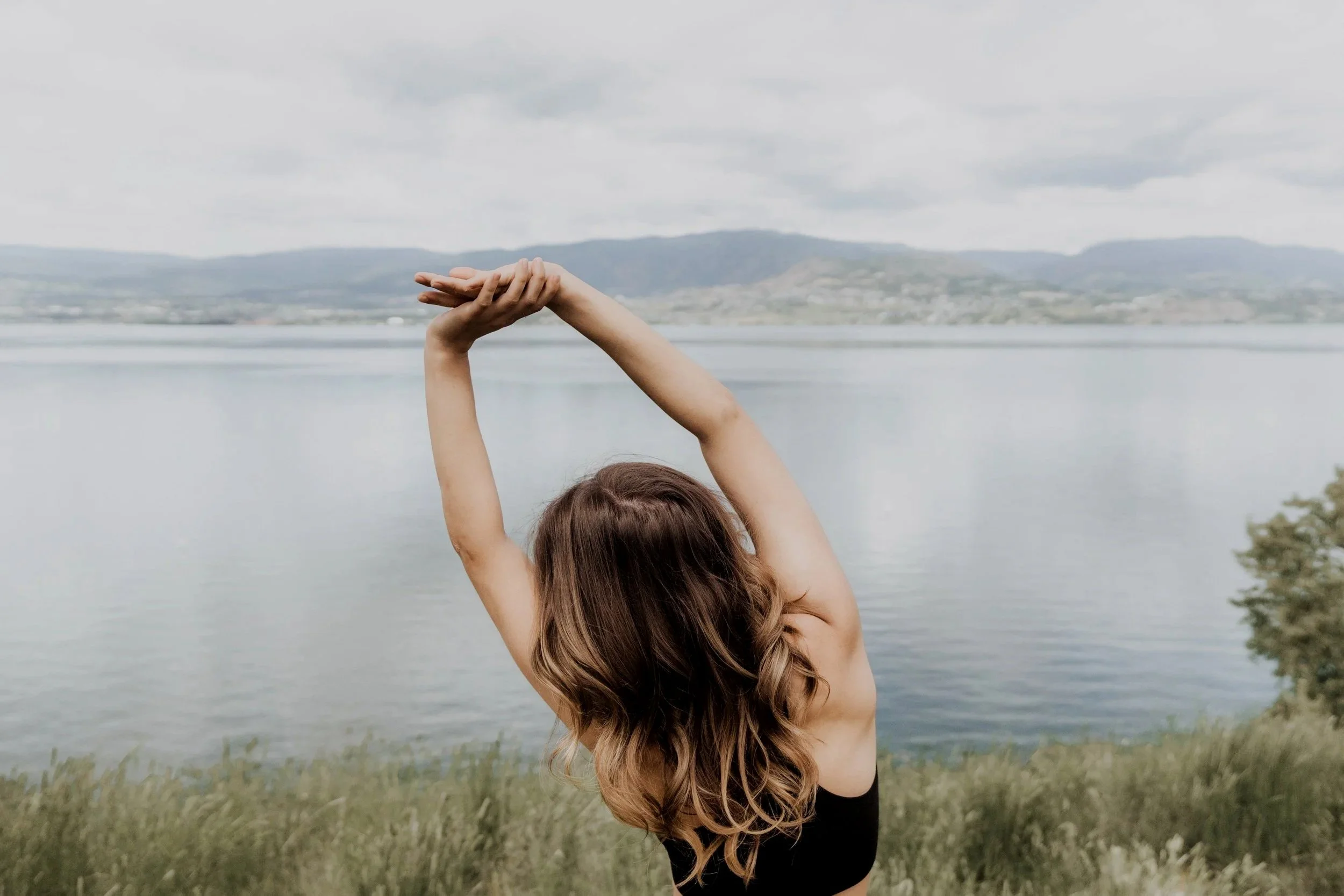 A woman with wavy brown hair practicing yoga outdoors by a lake with mountains in the background, stretching her arms above her in a side stretch.
