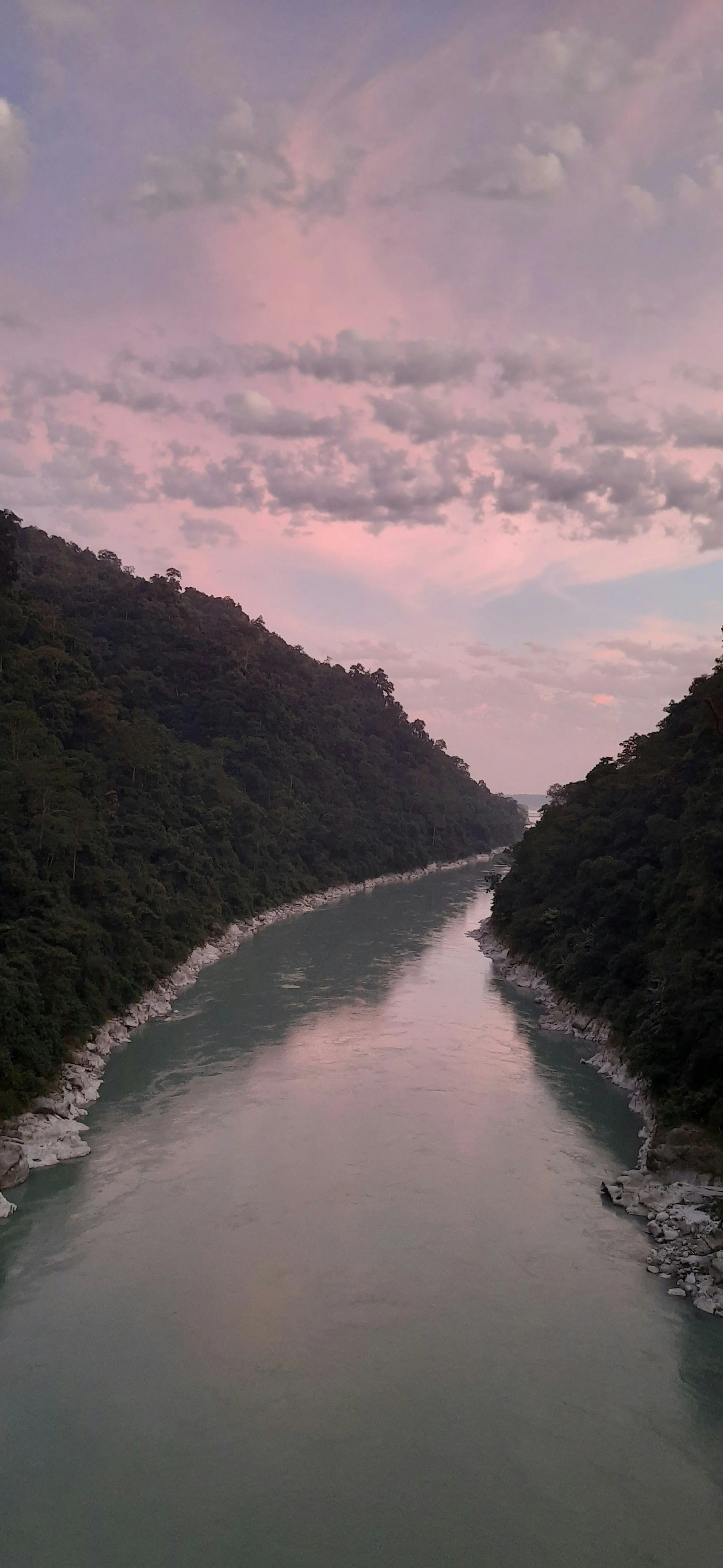 The Ganges River flowing between the forested foothills of the Himalayan mountains under a cloudy pink sky at sunset.