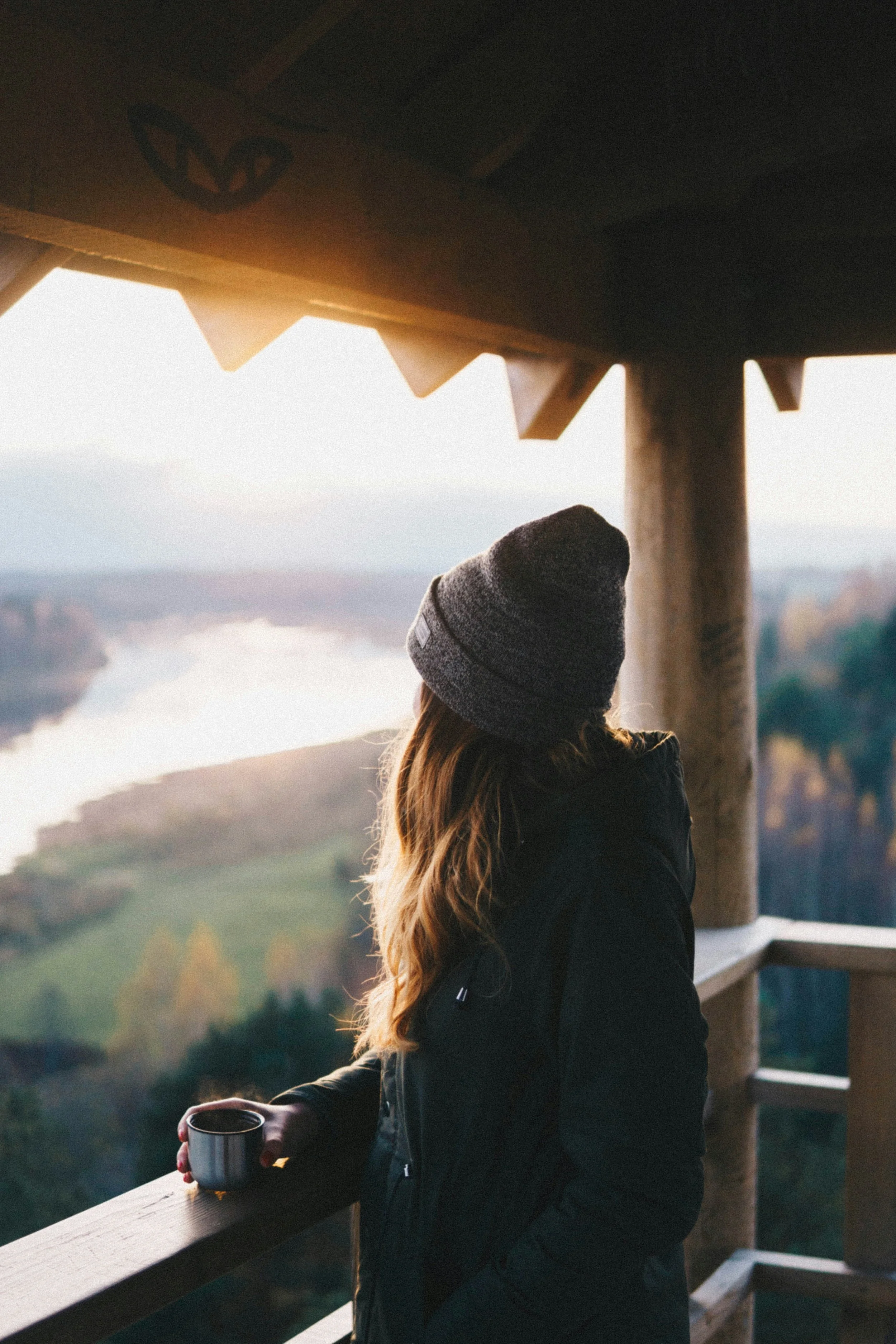 A woman in a gray beanie and black jacket calmly stands on a wooden balcony, holding a cup, overlooking a river and autumn-colored trees at sunrise or sunset.