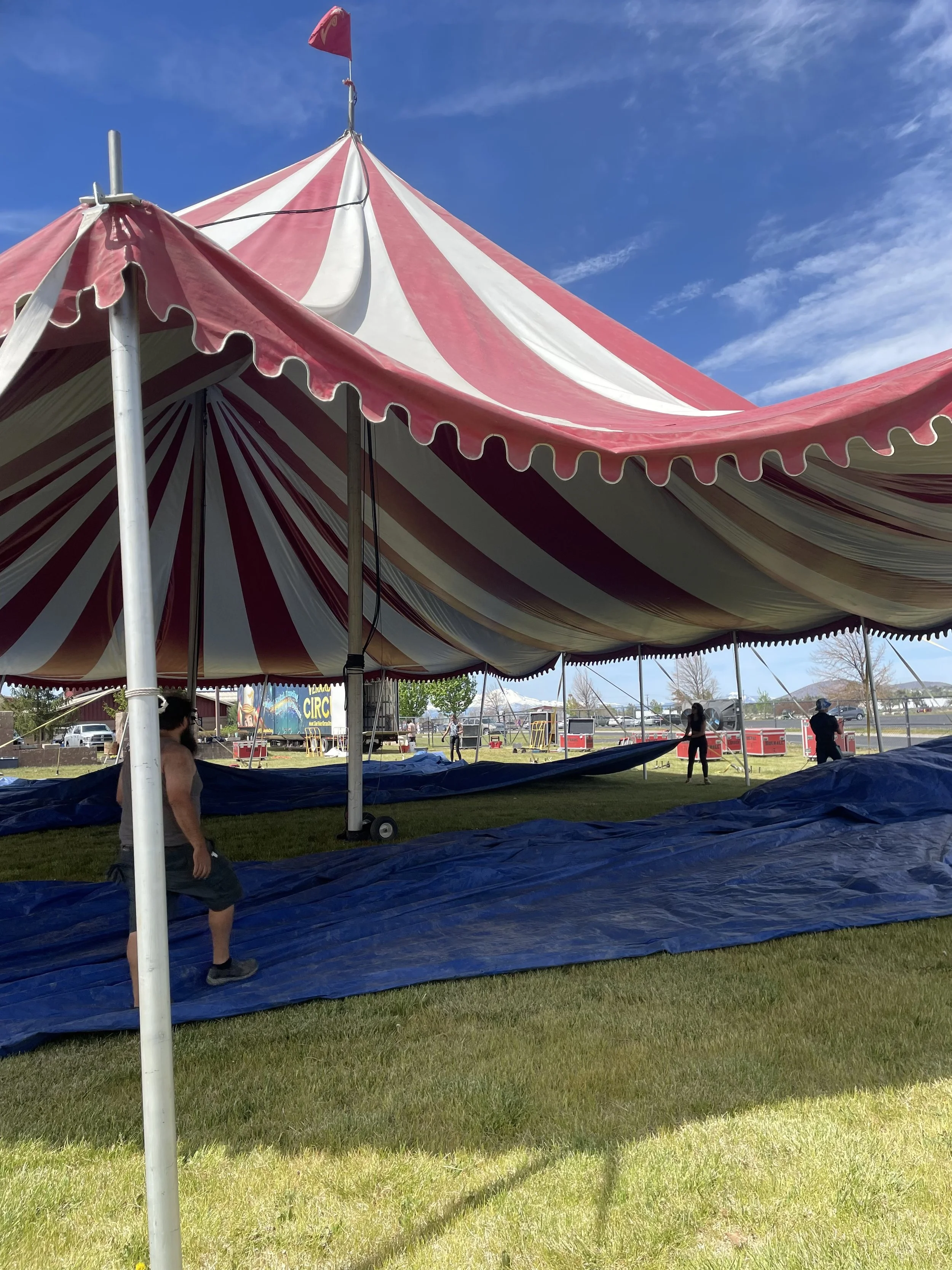 Circus tent being set up outdoors on a grassy field, with blue sky and a few clouds in the background.