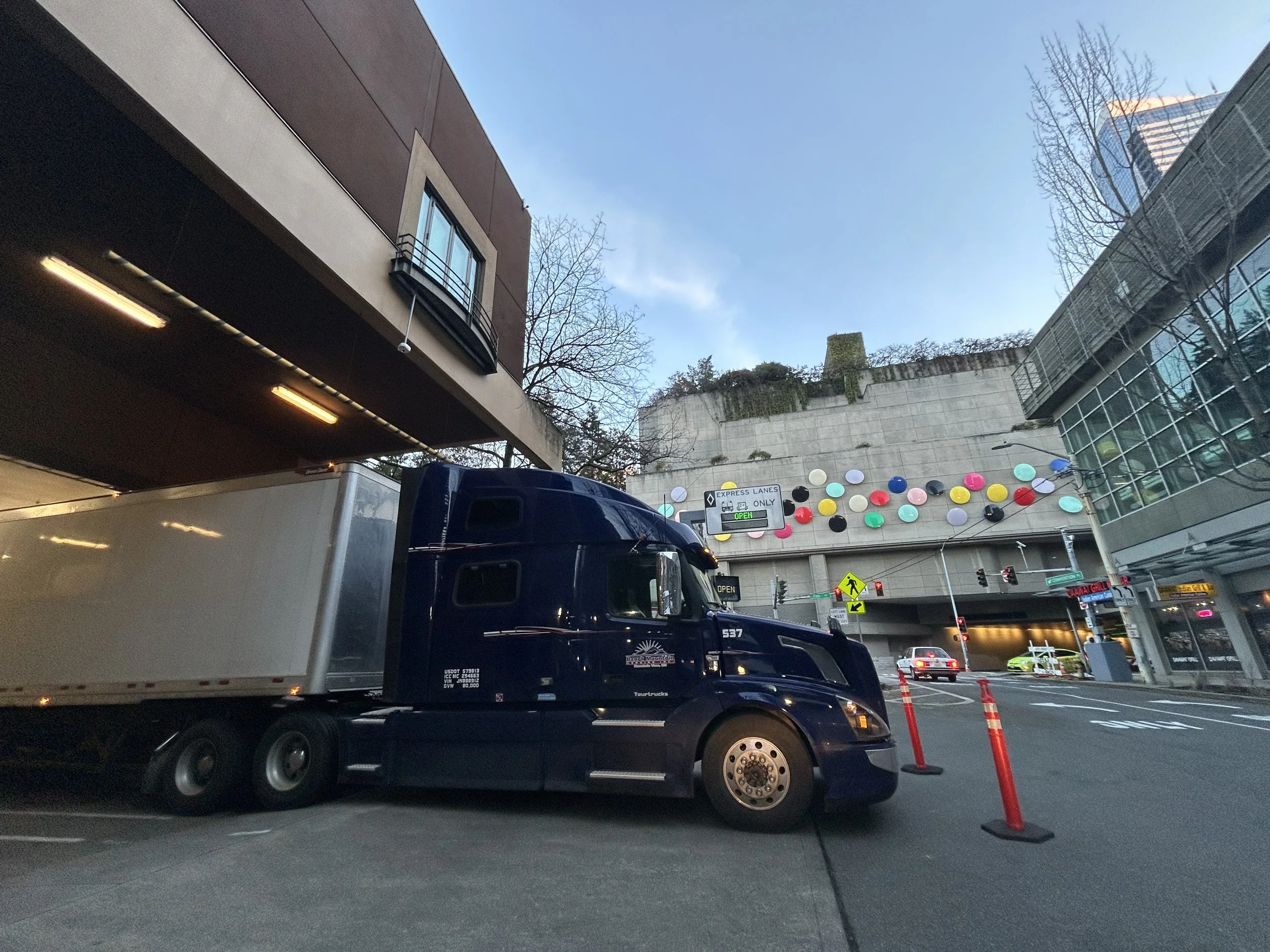 A large dark blue semi-truck with a long white trailer parked on city street. Buildings, traffic lights, colorful decorations on an overpass, and a clear sky are visible in the background.