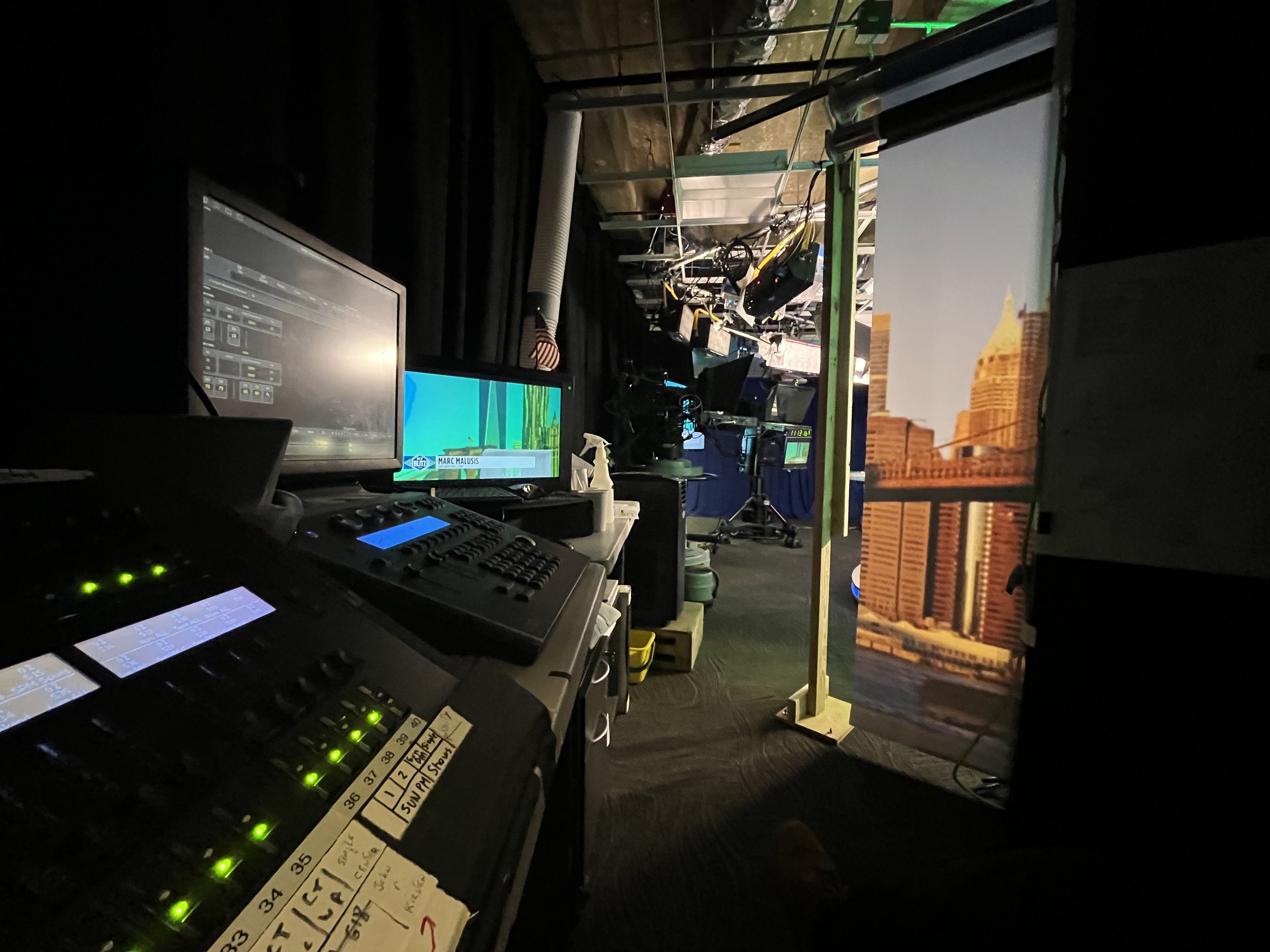 A behind-the-scenes view of a television studio control room with multiple monitors, equipment, and a view into the studio with a cityscape backdrop and stage lighting.