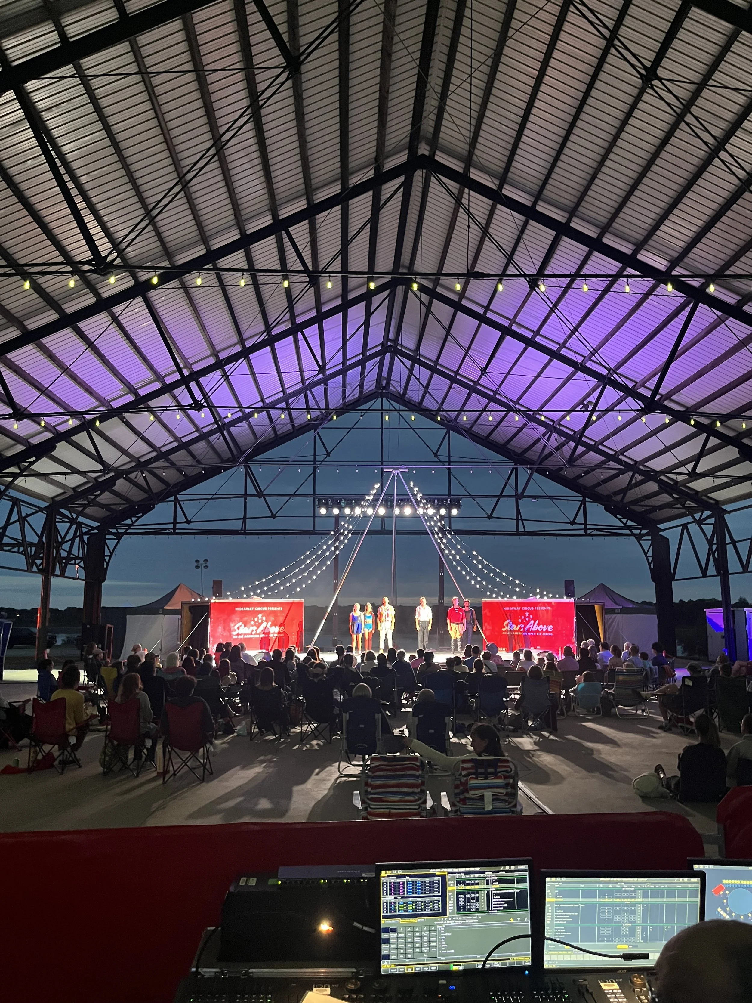Audience watching a circus performance under a large open-air roof with string lights and two large red screens on stage.