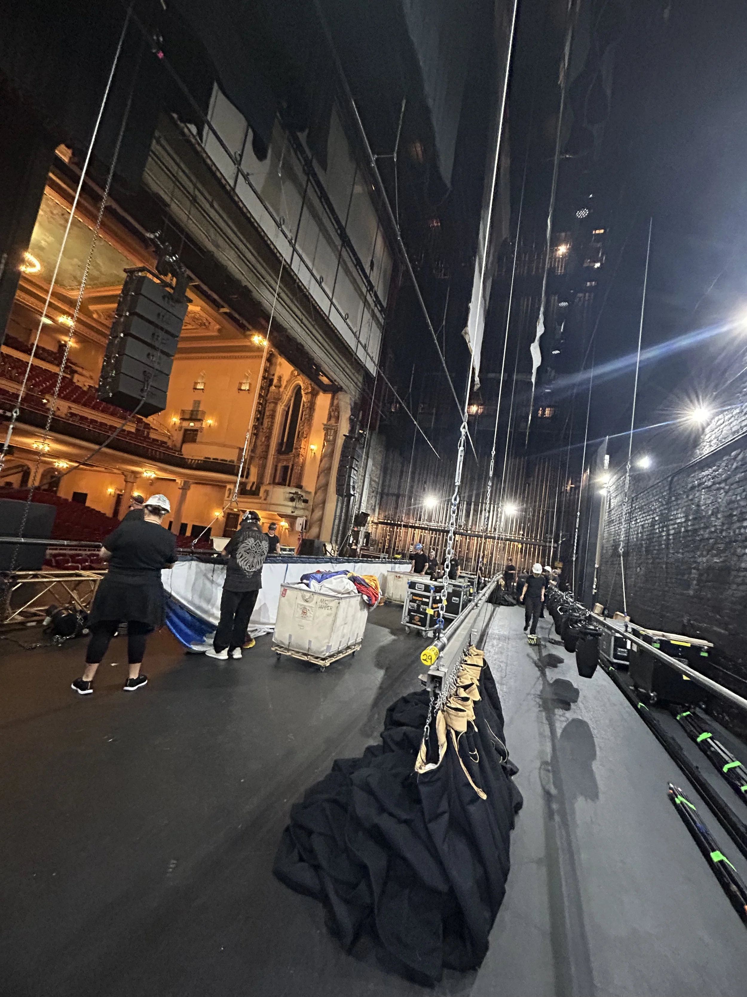 View of a stage setup in an empty theater with crew members preparing, black curtains along the sides, and seating area illuminated in the background.
