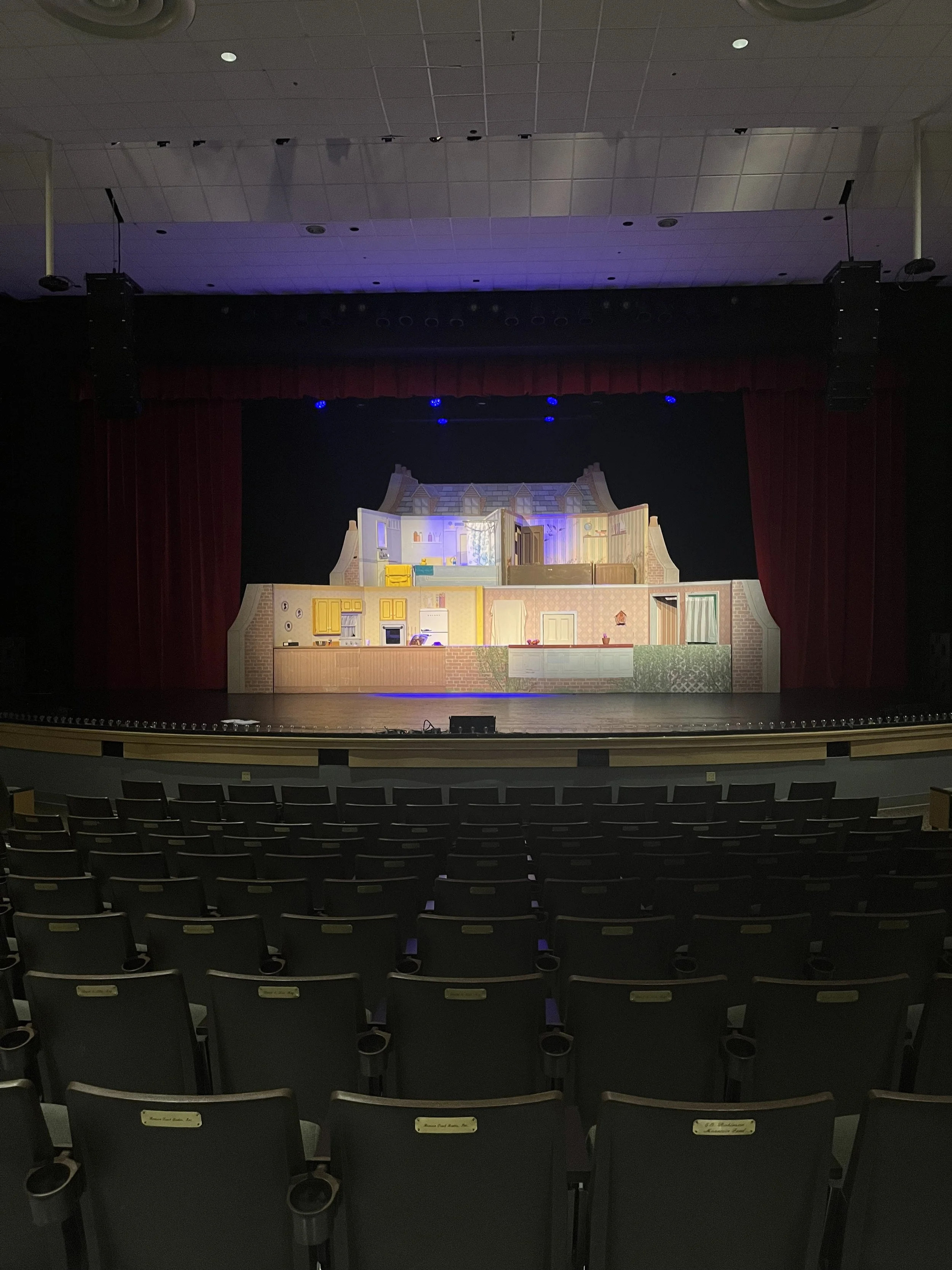 Empty theater auditorium with a stage set for a play or performance, featuring a large, detailed house facade backdrop with interior decor and furniture.